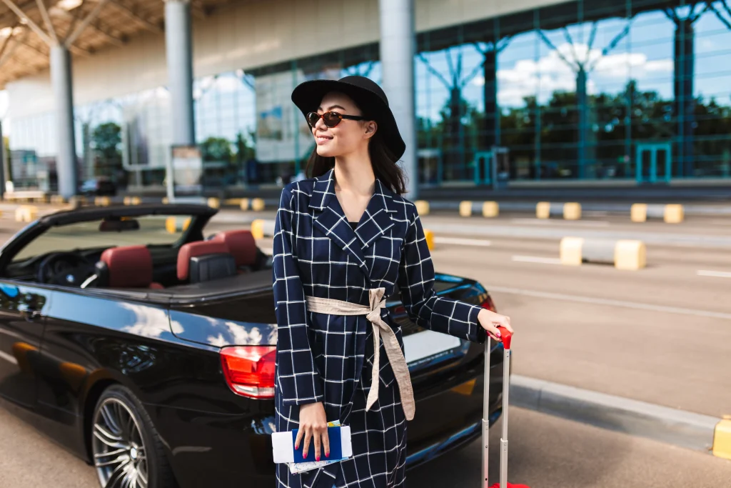hermosa chica sonriente con gafas de sol y sombrero negro con pasaporte con billete de avion mirando alegremente un lado cerca del aeropuerto con coche descapotable en el fondo