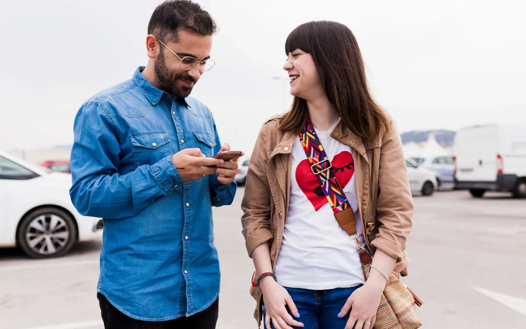 sonriente joven mirando hombre usando telefono celular en la calle
